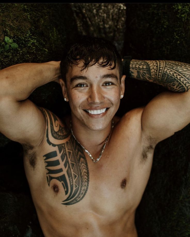 a shirtless man with tattoos on his arms and chest smiling at the camera while standing in front of a tree