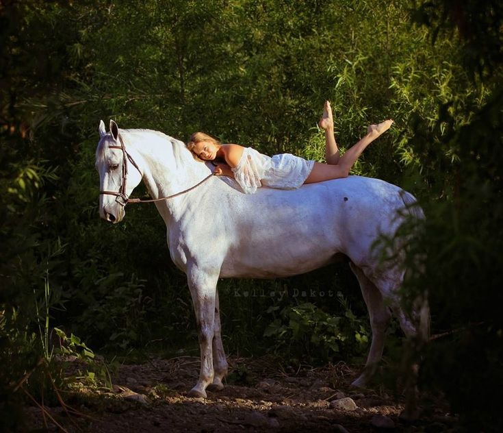 a woman sitting on top of a white horse in the woods with trees behind her