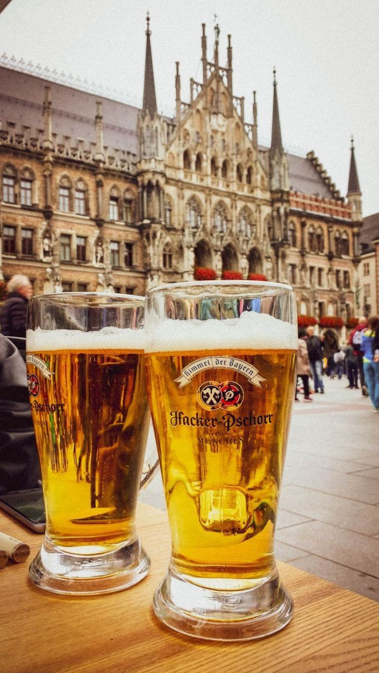 two beer glasses sitting on top of a wooden table in front of a castle like building