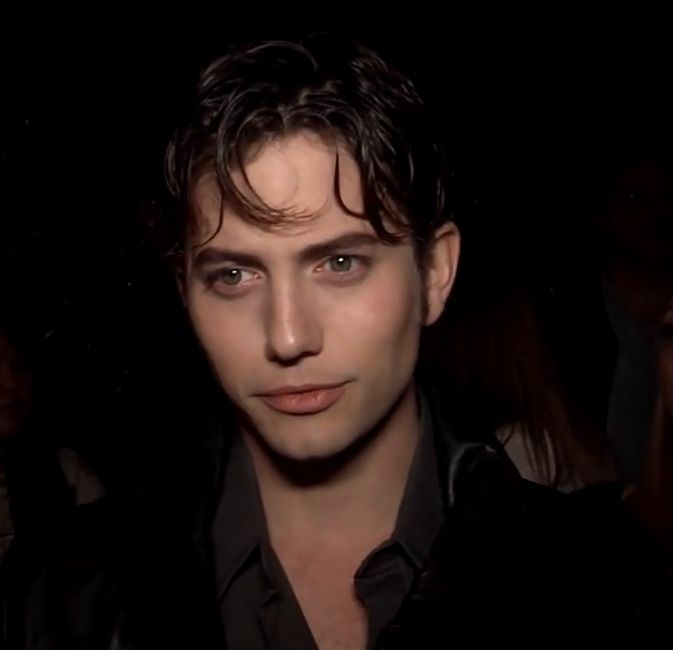 a man with dark hair and blue eyes is looking at the camera while people are standing around him