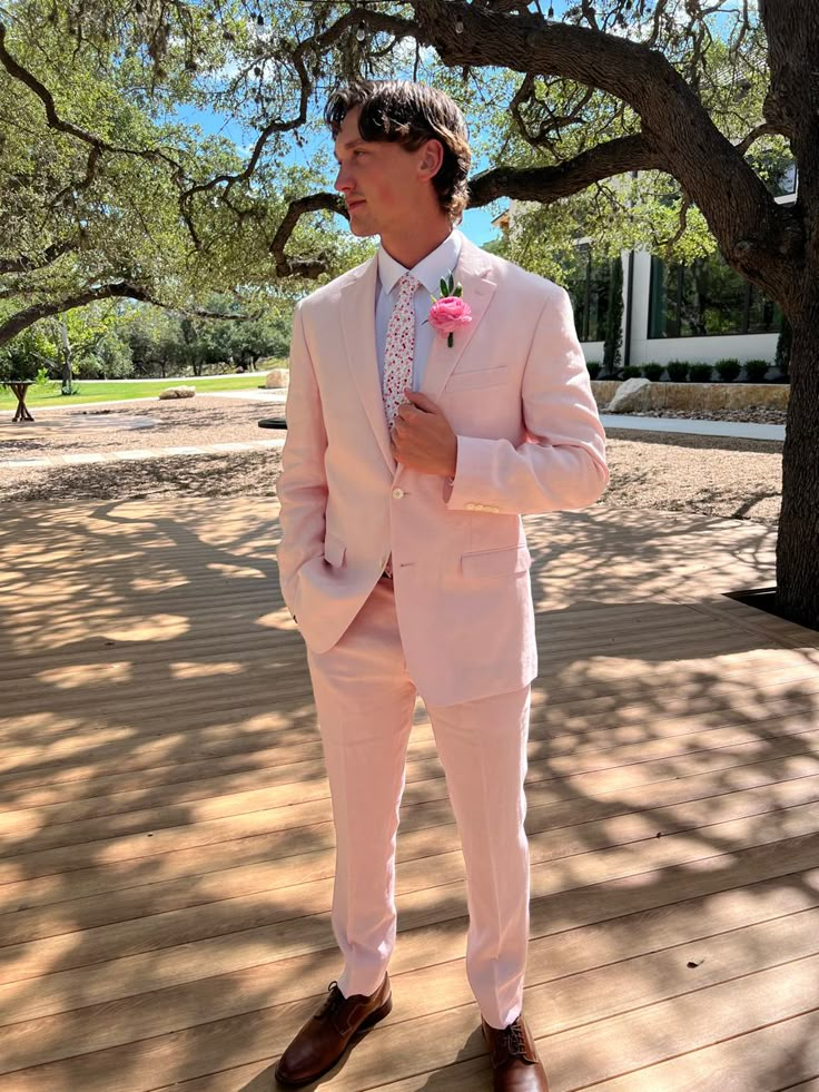 a man in a pink suit and tie standing on a wooden deck under a tree