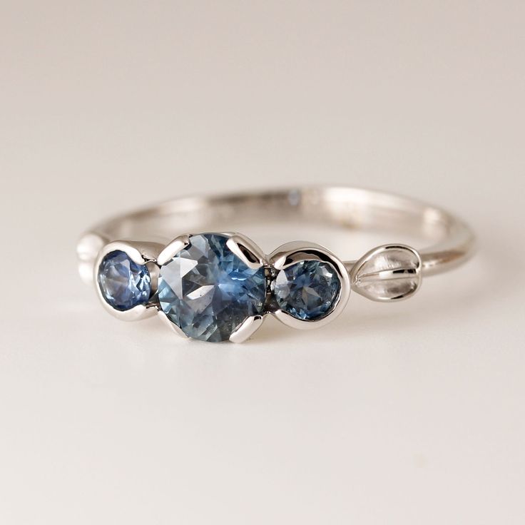 Montana blue sapphire ring, 5mm and two 3mm natural American sapphires. center sapphires have a carat weight of 0.68ct, the two side sapphires have a total weight of 0.26ct Set in a 14k white gold gold ring size 6 1/2. These lovely sapphires are partial bezel set accented bu a flowing leaf design. The shank is half round and tapers lightly. The ring is sizable, this design can also be made in 14k white or 14k yellow gold, platinum. This is my original design hand crafted in my northern Californi Blue Round Sapphire Ring With Rose Cut Diamonds, Three Stone Blue Topaz Sapphire Promise Ring, Blue Sapphire Ring With Rose Cut Diamonds For Anniversary, Anniversary Blue Sapphire Ring With Rose Cut Diamonds, Blue Sapphire Ring In 14k Gold With Brilliant Cut, Blue Brilliant Cut Sapphire Ring In 14k Gold, Fine Jewelry Three-stone Sapphire Ring, Sapphire Promise Ring With Rose Cut Diamonds, Blue Sapphire Three-stone Birthstone Ring