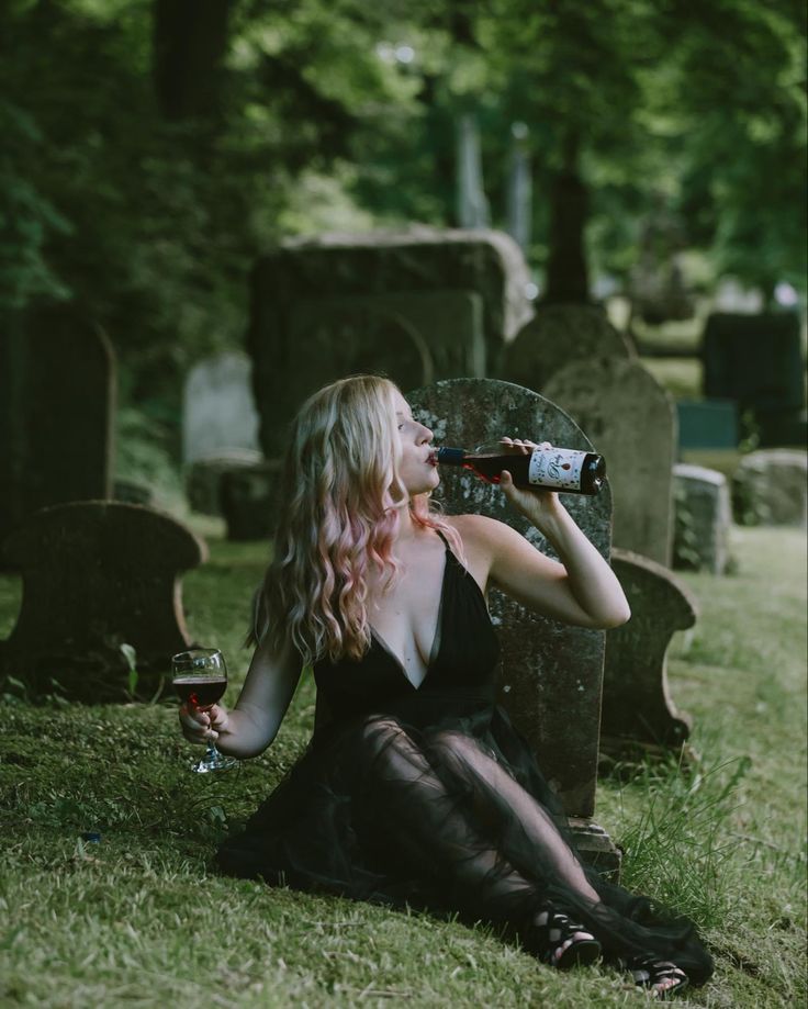 a woman sitting on the ground drinking from a bottle and holding a glass in her hand