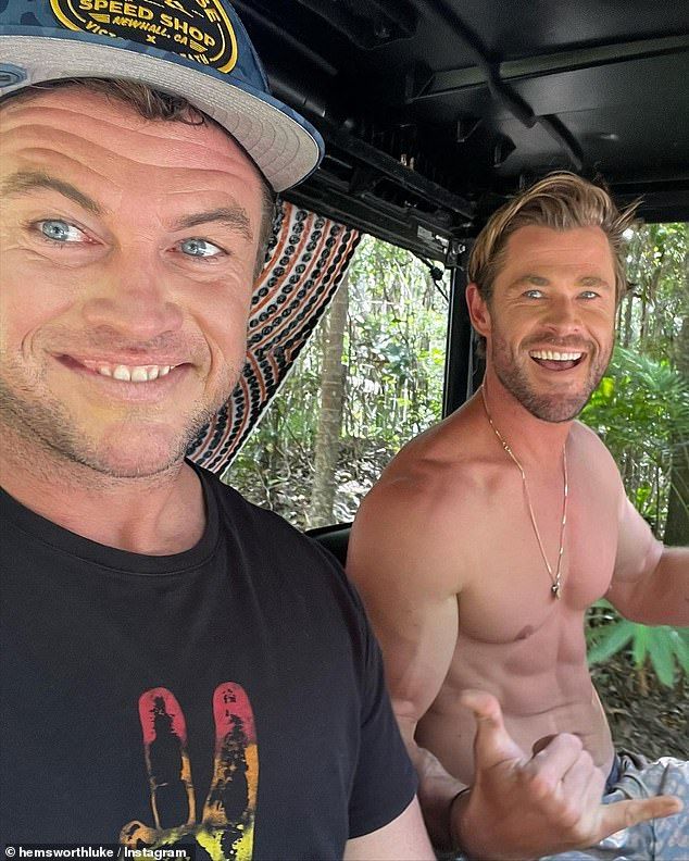 two shirtless men sitting in the back of a truck with trees and bushes behind them