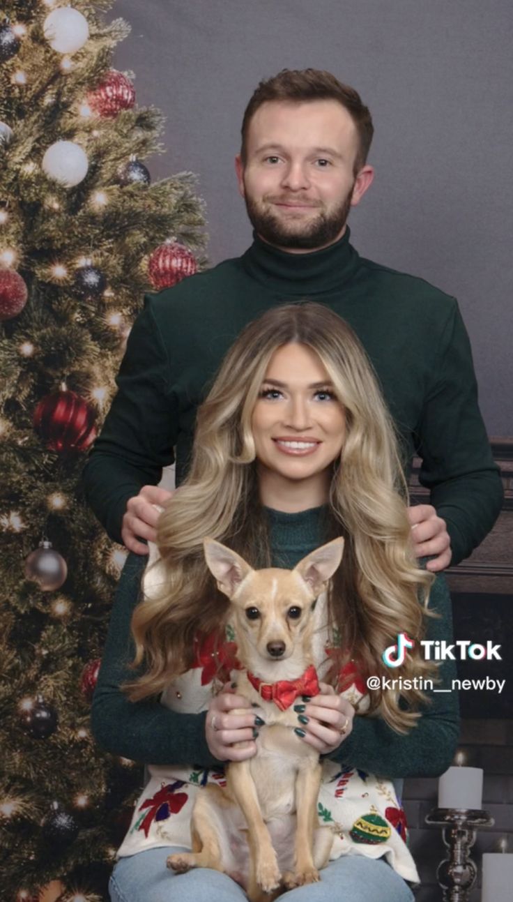 a man and woman sitting in front of a christmas tree with their dog on his lap