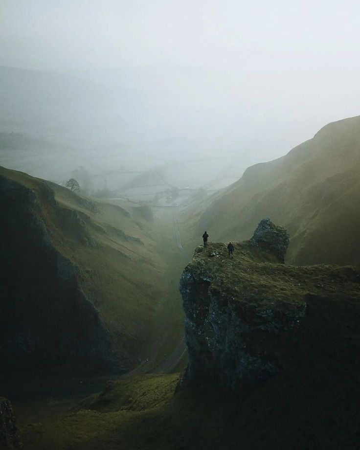 two people are standing on the edge of a cliff in the foggy hills above them