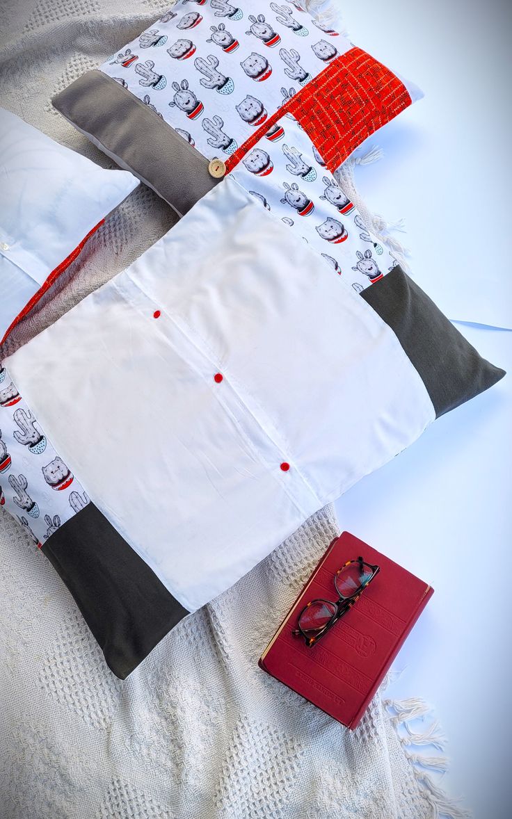 three pillows and a red book on a white bed spread with the covers folded down