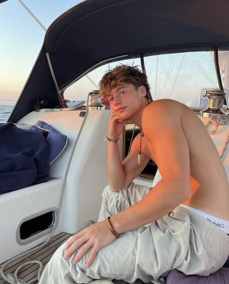 a shirtless man sitting on the bow of a sailboat while talking on his cell phone