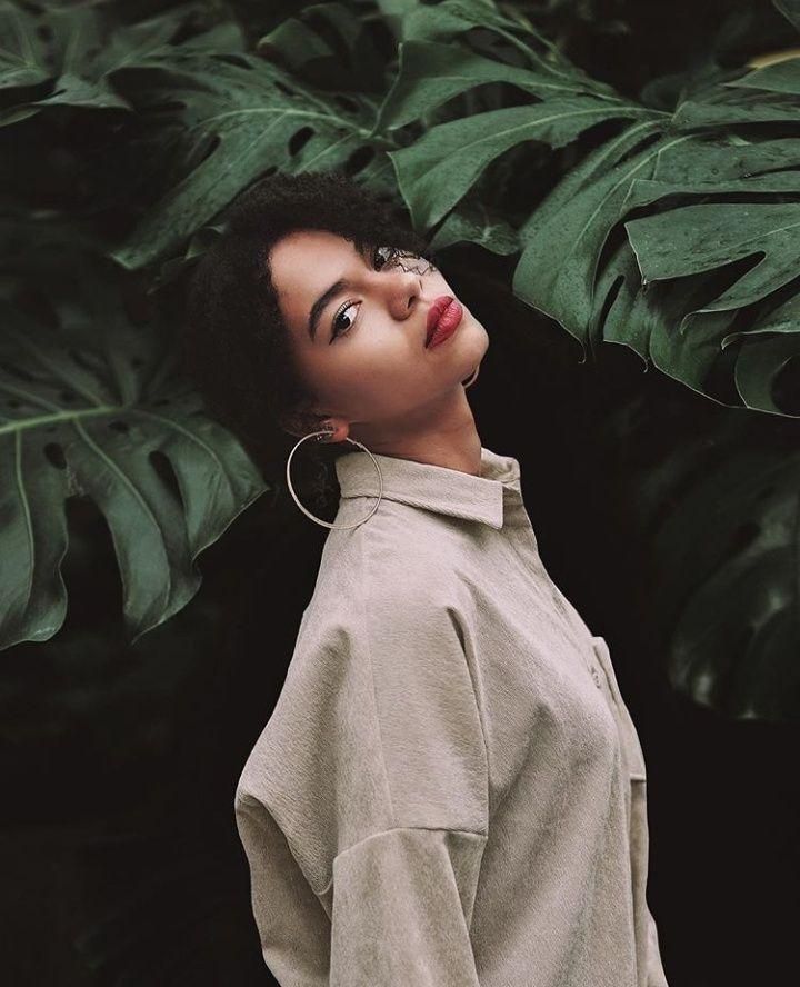 a woman is standing in front of some plants and looking up at the sky with her eyes closed