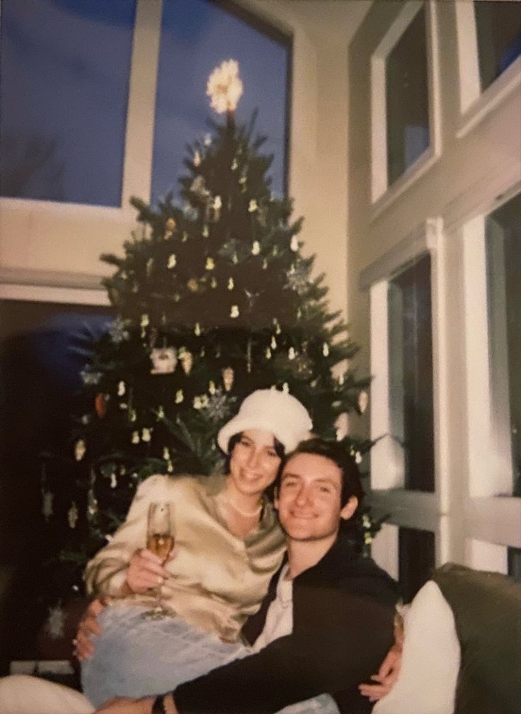 a man and woman sitting next to a christmas tree with a glass of wine in front of them