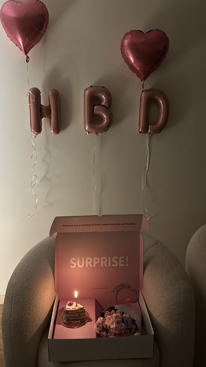 a birthday cake in a box with balloons on the wall behind it that says surprise