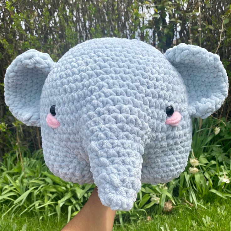 a crocheted elephant head is shown in front of some grass and bushes with pink eyes