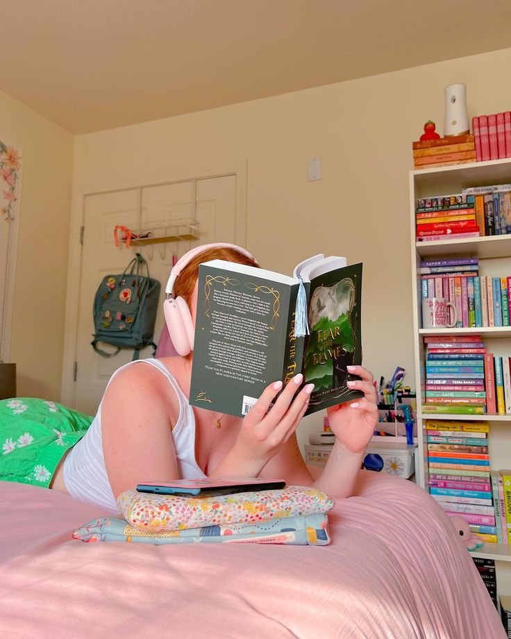 a woman laying in bed reading a book with headphones on her ears and holding an open book