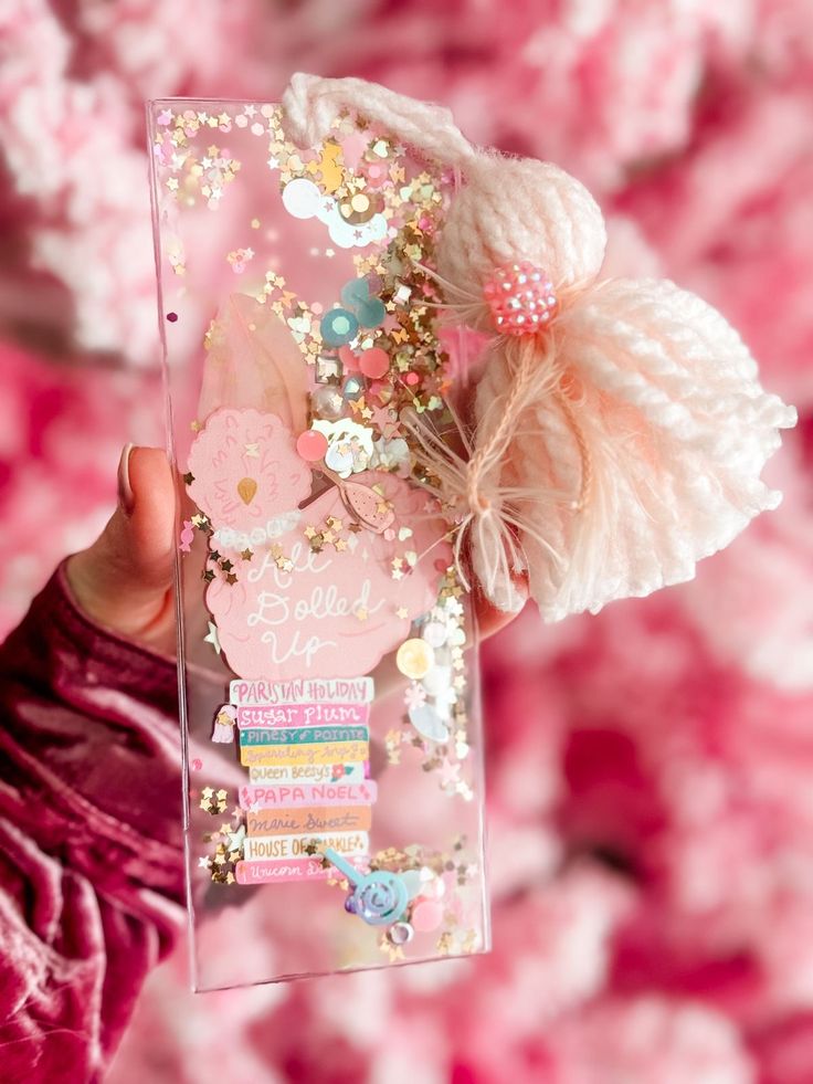 a person holding up a clear case with flowers and beads on the inside, in front of a pink background