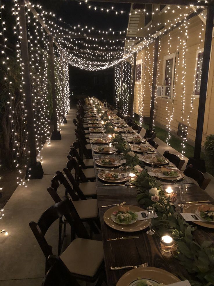 a long table set with plates and place settings for dinner outside at night, lit by fairy lights