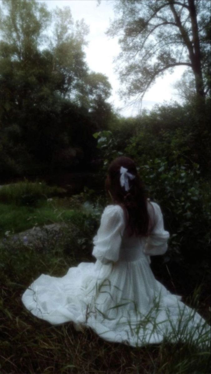 a woman in a white dress is sitting on the grass and looking into the distance