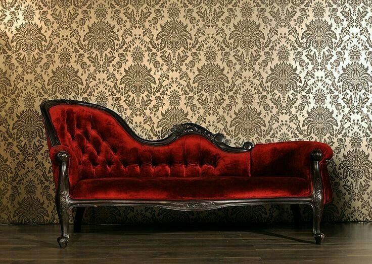 a red velvet couch sitting in front of a wallpapered wall with damask