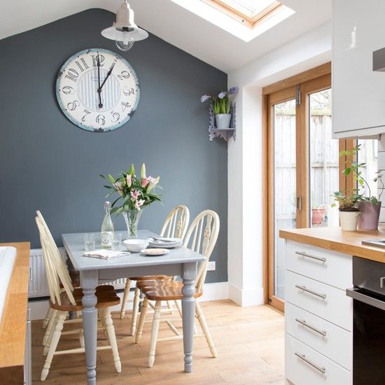 an image of a kitchen setting with blue walls and white furniture in the background, there is a large clock on the wall above the table