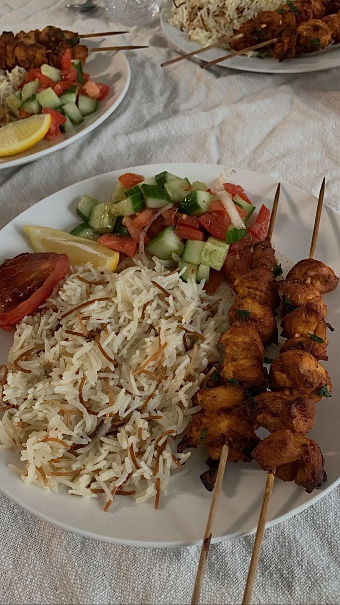 two plates filled with different types of food on skewers next to rice and cucumbers