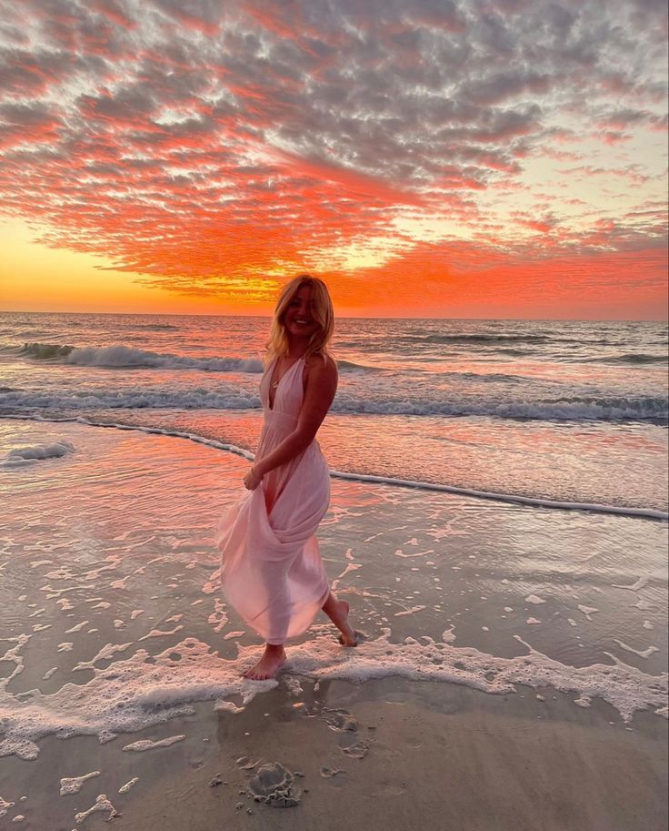 a woman in a white dress walking on the beach at sunset with her feet in the water