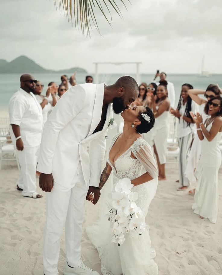 a bride and groom kissing on the beach in front of their wedding party, surrounded by other people