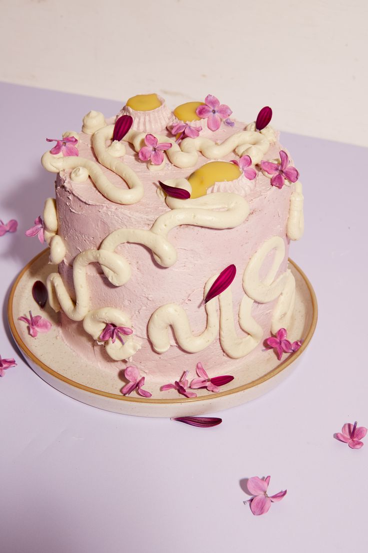 a pink cake with white frosting and flowers on it sitting on a table next to confetti