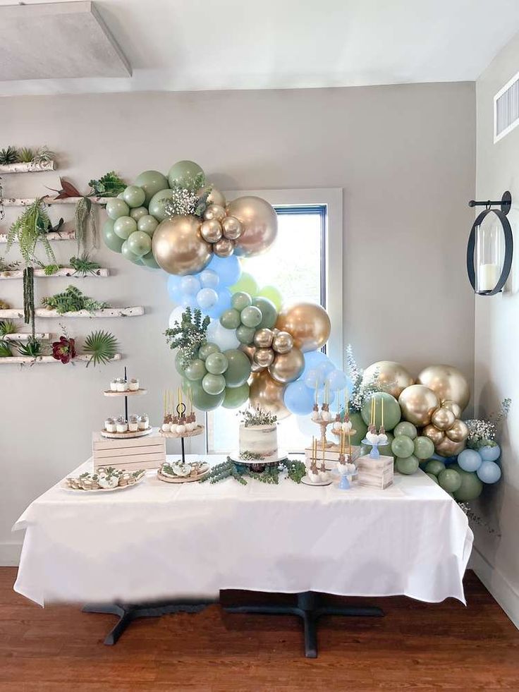 a white table topped with lots of balloons and cake next to a window covered in greenery