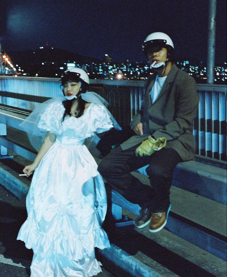 a man and woman dressed in wedding attire sitting on a bench at night with city lights behind them