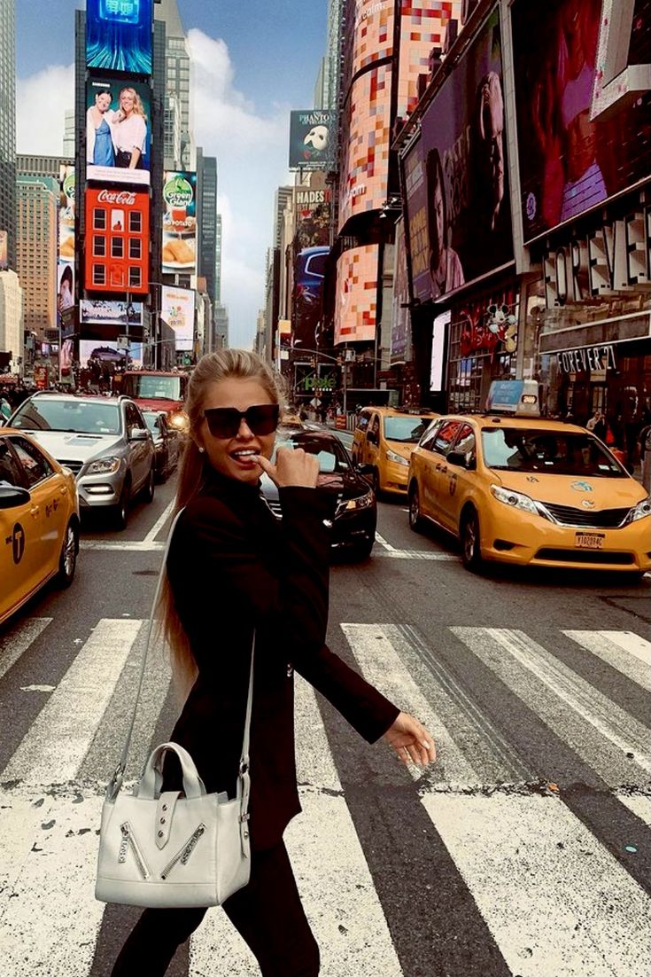 a woman walking across a cross walk in the middle of a busy city with taxi cabs