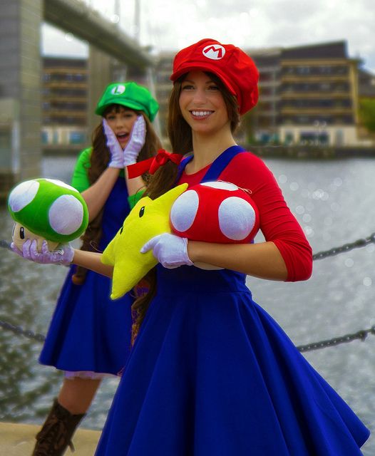 two women dressed in mario and luigi bros costumes, one holding a stuffed animal toy