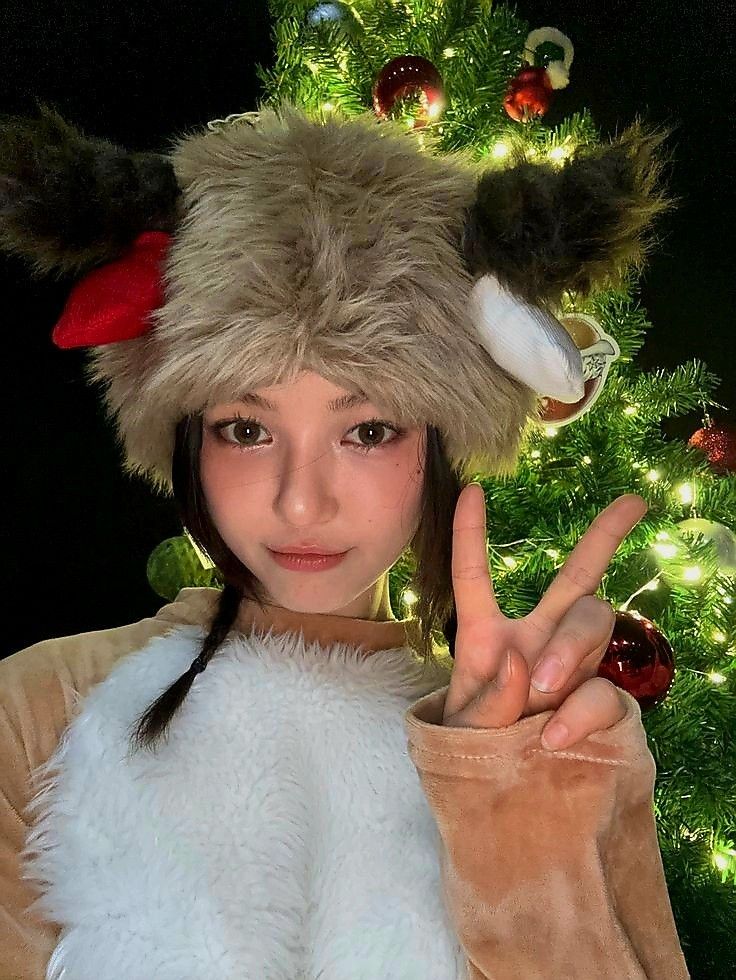 a woman wearing a furry hat making the peace sign with her hand while standing in front of a christmas tree