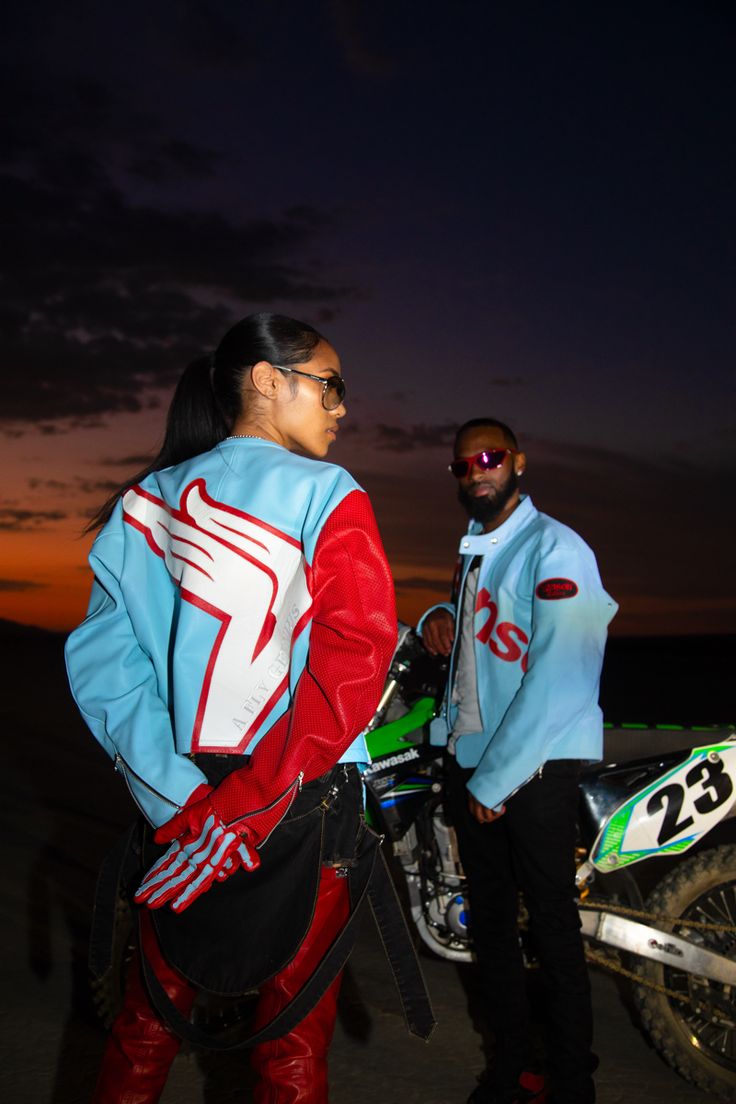 two people standing next to each other in front of a dirt bike at night with the sun setting behind them