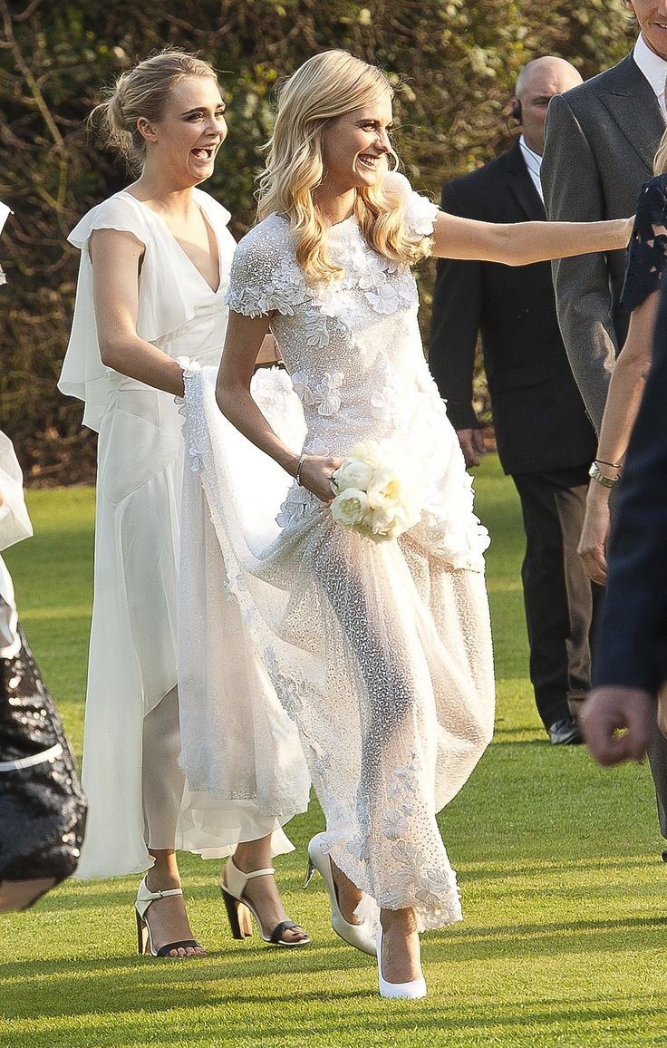 two women in white dresses are walking through the grass with one woman holding her hand out