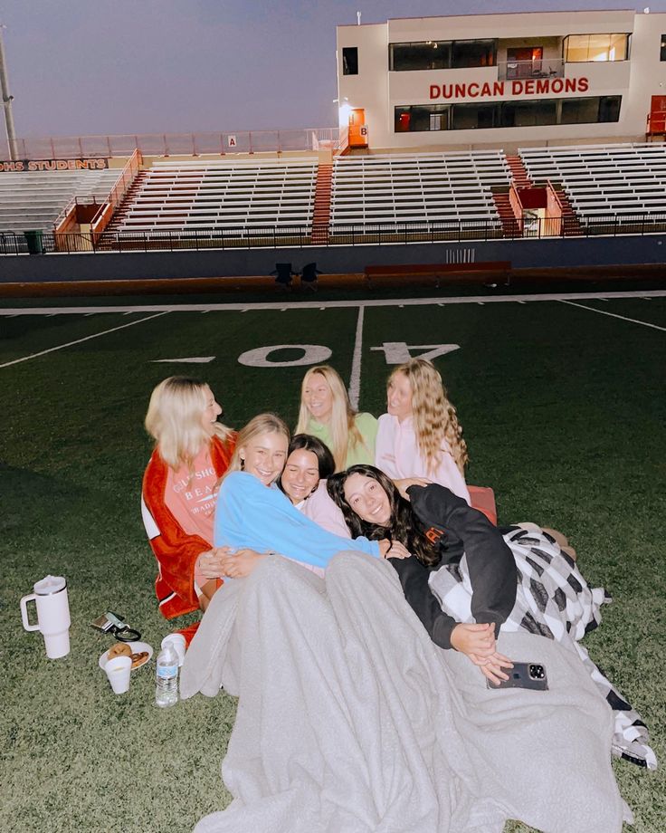 four women sitting on the ground in front of an empty football field at night time