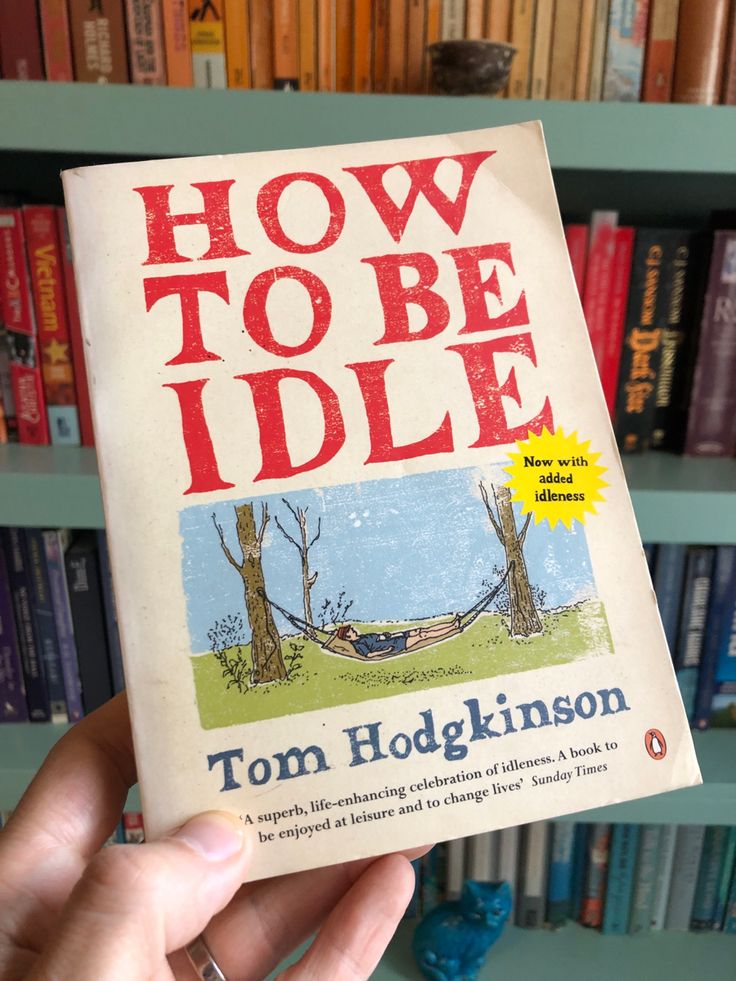 the book how to be idle by tom hodkinson is held in front of a bookshelf