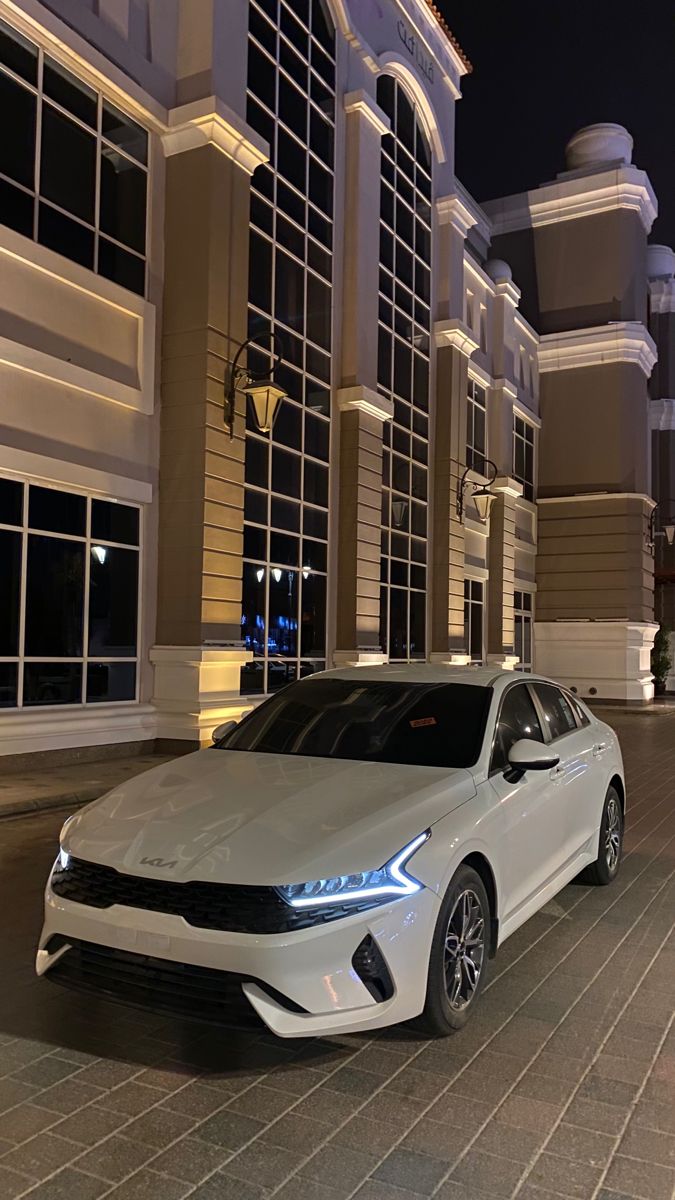 a white car is parked in front of a building at night with its lights on