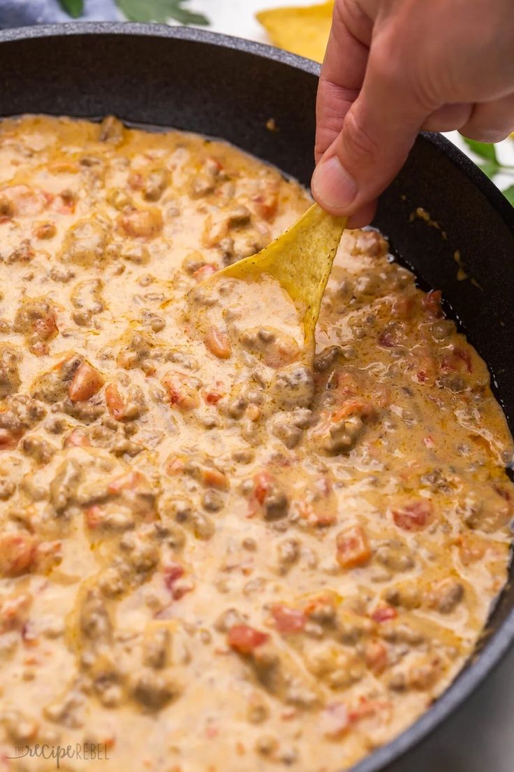 a person dipping a tortilla chip into a skillet filled with cheese and meat