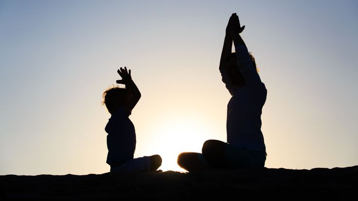 two people sitting on the ground with their hands up in the air while the sun is setting