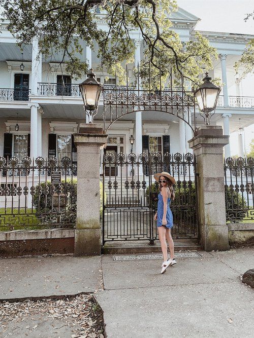 a woman standing in front of a gate with a hat on her head and wearing a blue dress