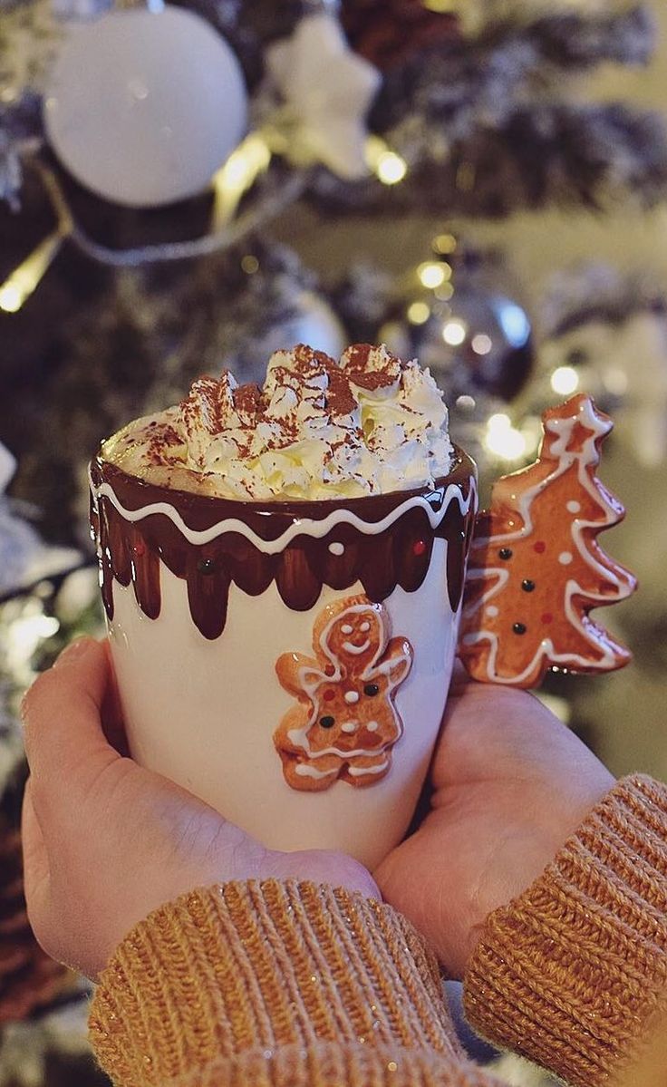 a hand holding a cup filled with hot chocolate and marshmallows next to a christmas tree