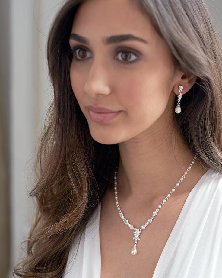 a woman wearing a necklace and earrings with pearls on the bottom, standing in front of a window