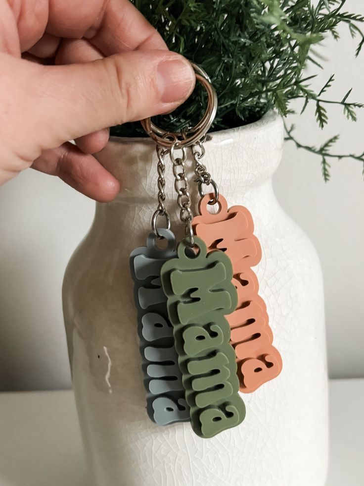 a hand holding a keychain with the word grandma on it, in front of a potted plant