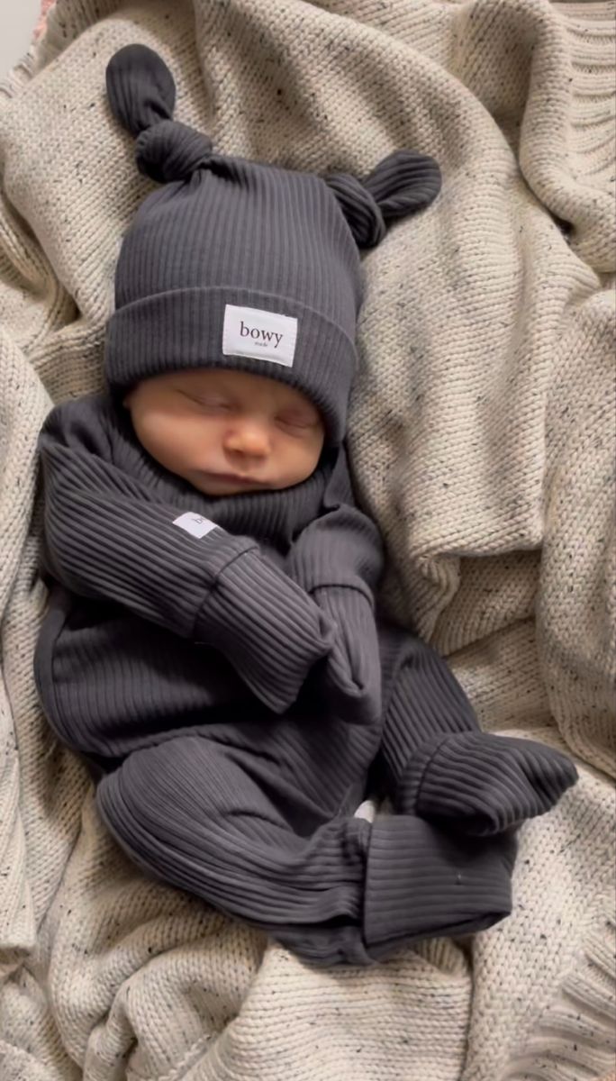 a baby wearing a bear hat and grey sweater laying on a pile of knitted blankets