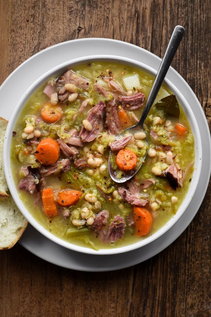 a bowl of soup with meat, beans and carrots on a plate next to a slice of bread