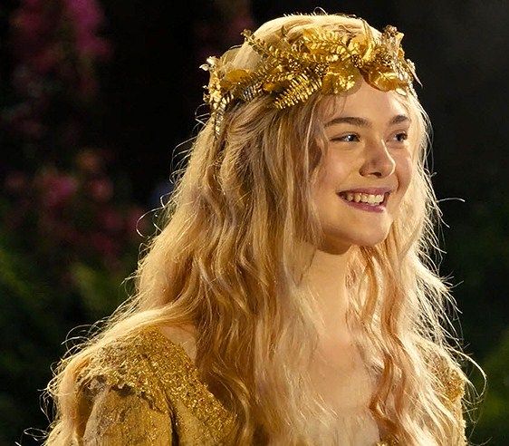 a woman with long blonde hair wearing a gold crown and smiling at the camera while standing in front of flowers