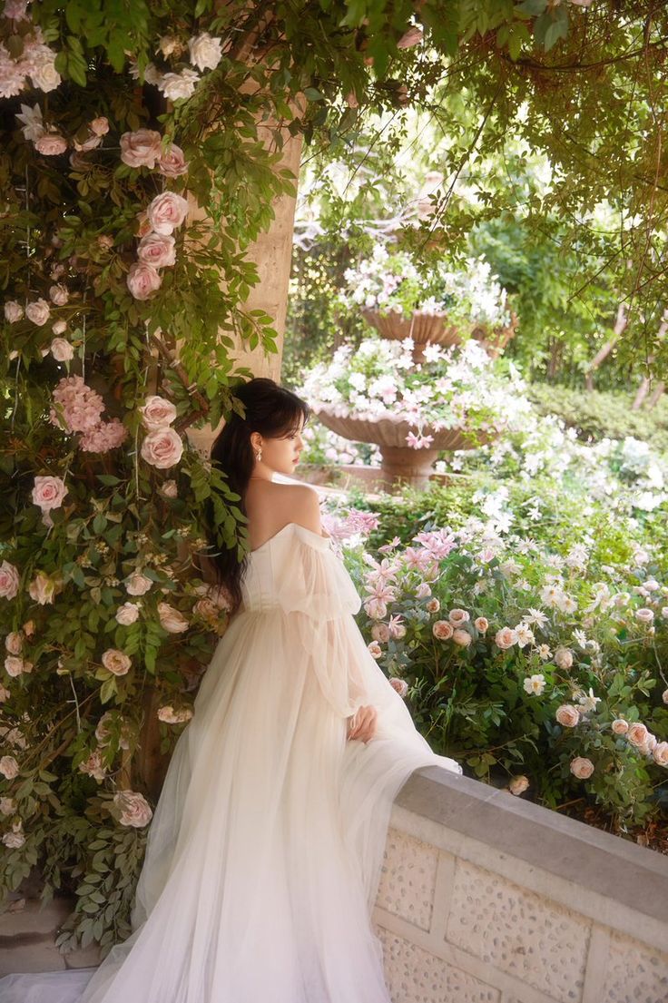 a woman in a white dress leaning against a wall with flowers on it and greenery behind her