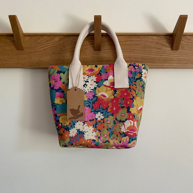 "This pretty mini-tote is made from Liberty fabric 'Flowers of Thorpe' in Summer Bloom. The dense small floral design is printed on a soft linen-cotton blend fabric. Lined with a durable gold linen blend fabric with an interior slip pocket, removable insert which fits neatly into the bottom of the bag and strong cord-centre ivory handles.  It also features our exclusive laser-etched hardwood Jenney Wren label on the inside pocket. Sizing: This bag has a base approximately 3.5\" (19cm) x 4.7\" (12cm) and is 8.6\" (22cm) deep. The handles peak approximately 4.7\" (12cm) above the top of the bag." Eco-friendly Floral Canvas Bag For Everyday Use, Casual Rectangular Canvas Bag With Floral Print, Spring Cotton Bags With Double Handle, Summer Everyday Canvas Bag, Spring Fabric Tote Shoulder Bag, Summer Floral Print Canvas Shoulder Bag, Travel Cotton Bag With Floral Print, Summer Cotton Canvas Bag With Handles, Cotton Travel Bag With Floral Print