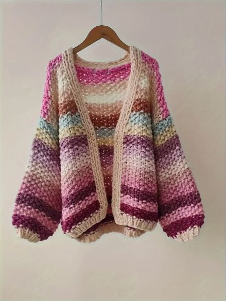 a knitted sweater hanging on a hanger in front of a white wall with a pink, purple and green striped cardigan