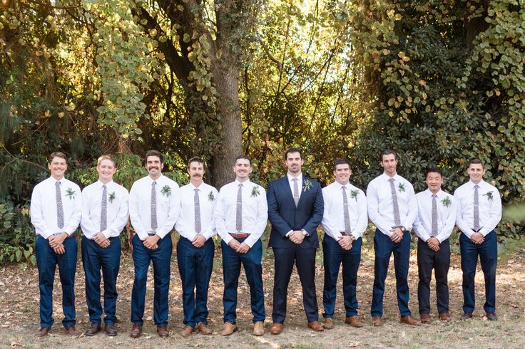 a group of men standing next to each other in front of trees and bushes with ties on