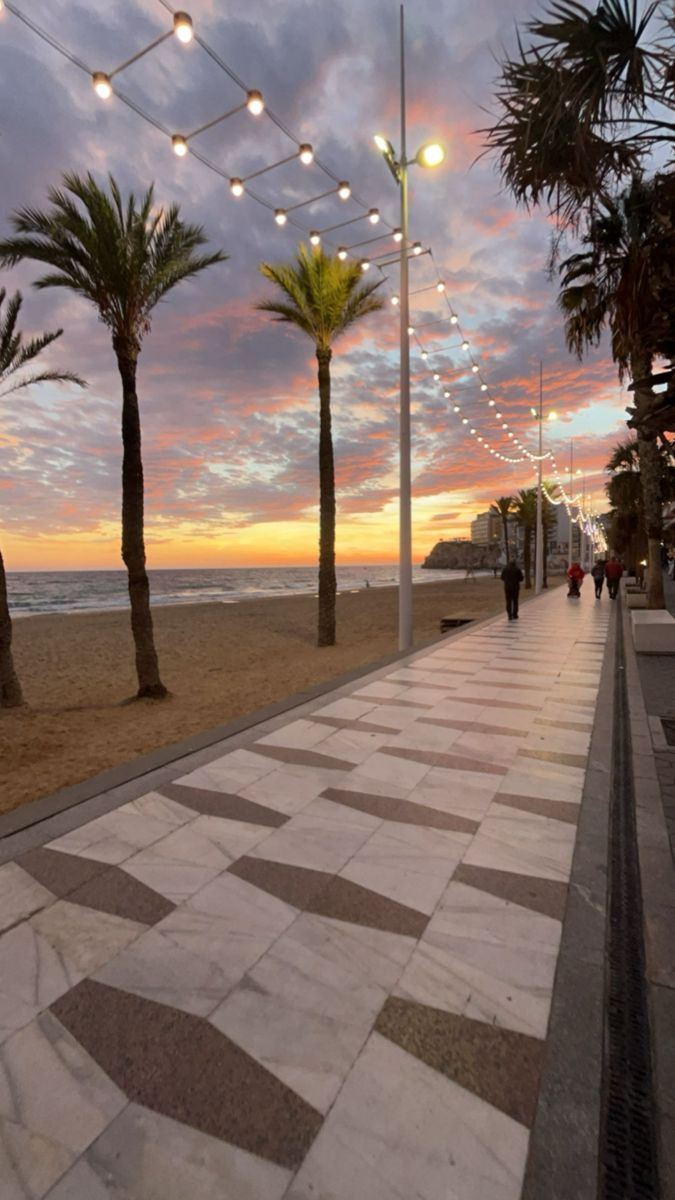 people walking on the beach at sunset with palm trees in the foreground and lights strung across the walkway